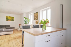 Een moderne woonkamer met een houten vloer, een grijze bank, een eettafel met stoelen, en een keuken met een wit aanrecht en lades.