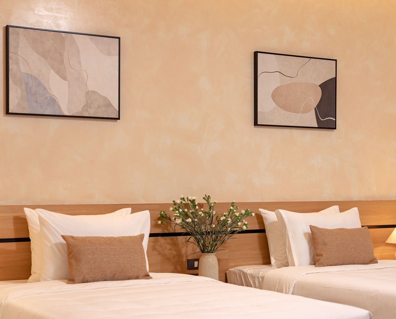 A hotel room with two beds, beige walls, and abstract art on the wall.