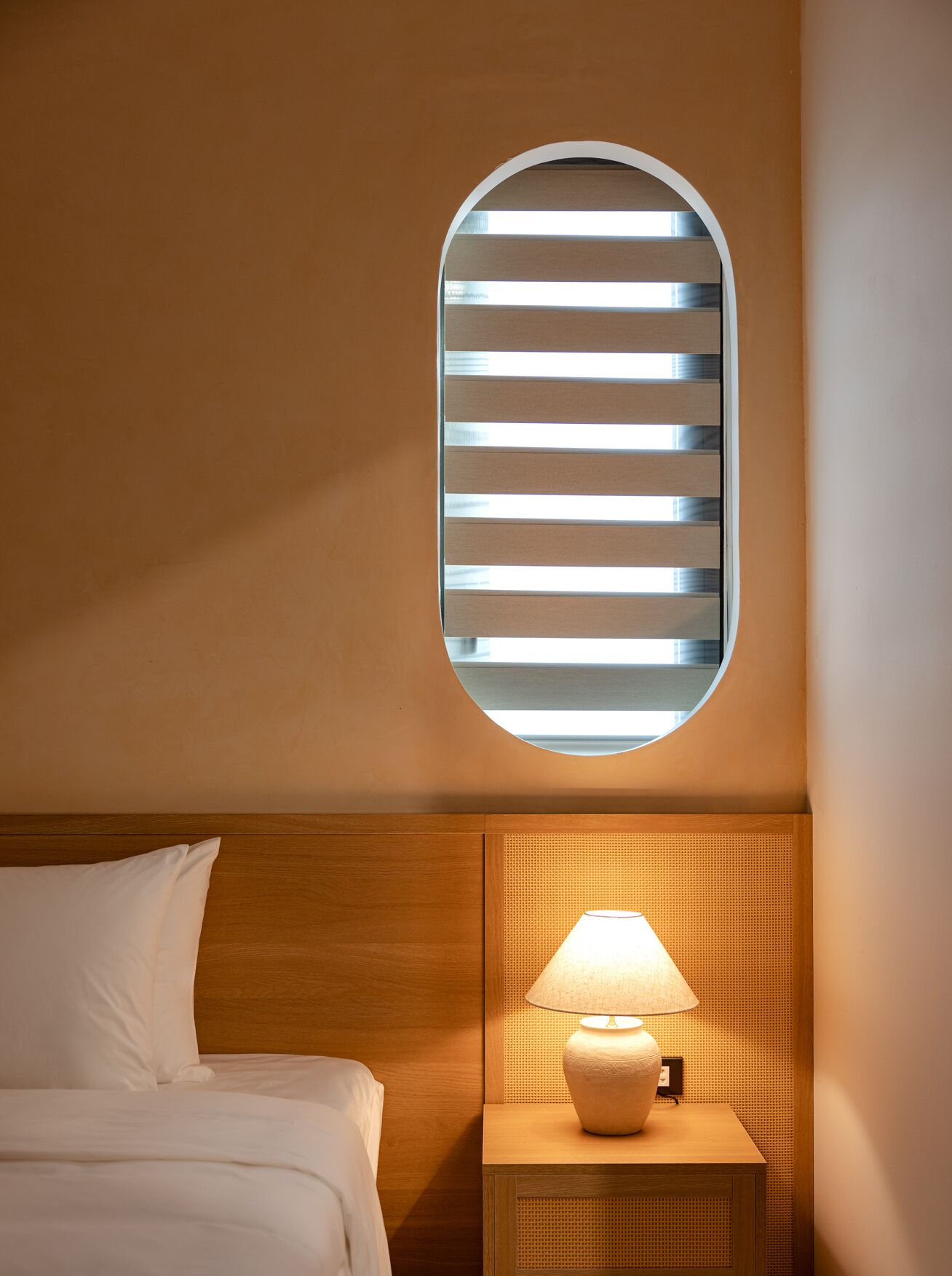 Una habitación de hotel con cama, una mesita de noche con lámpara y un ventanal circular con persianas horizontales.
