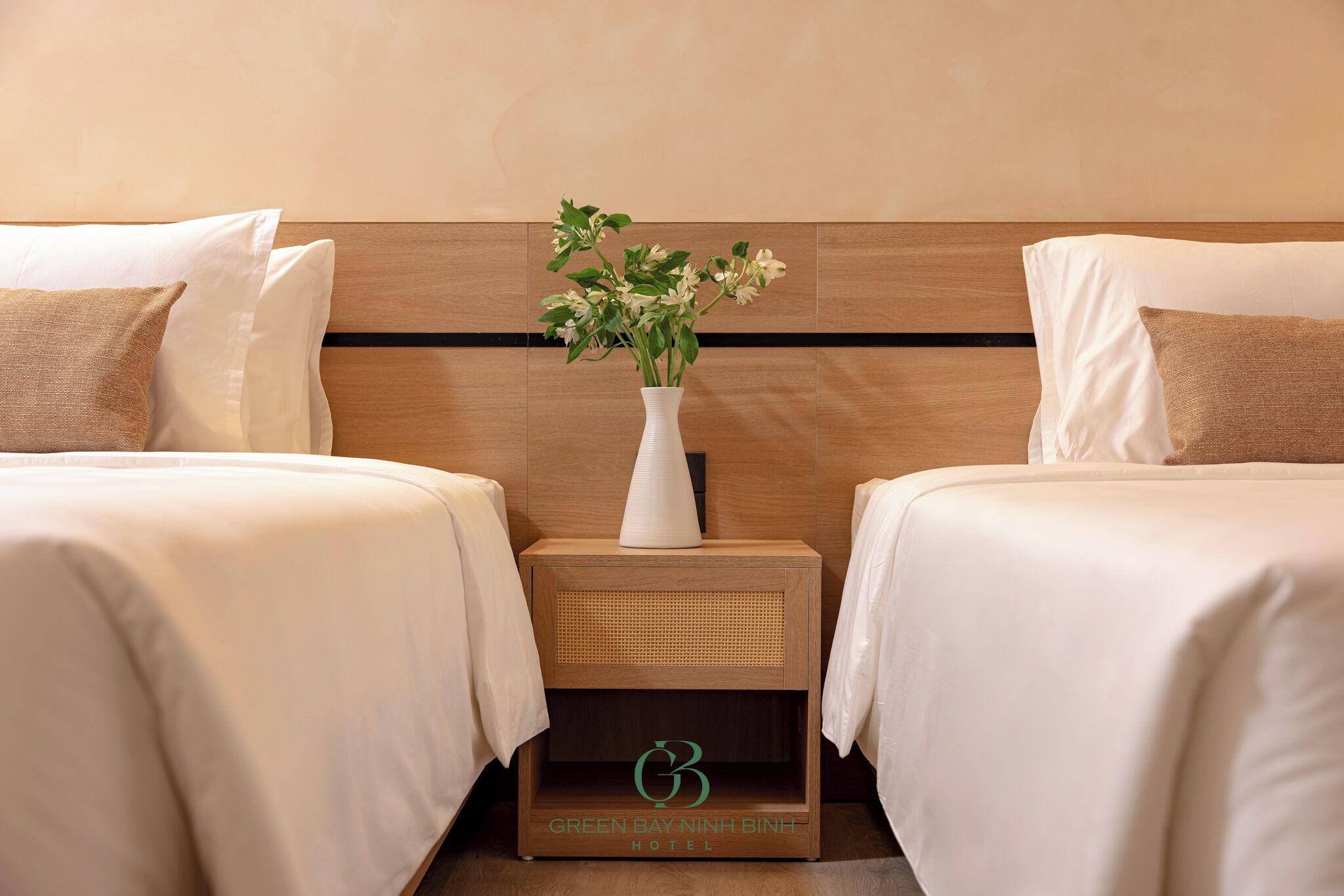 Habitación de hotel con dos camas, una mesita de noche con un jarrón de flores y un logotipo en la mesita.