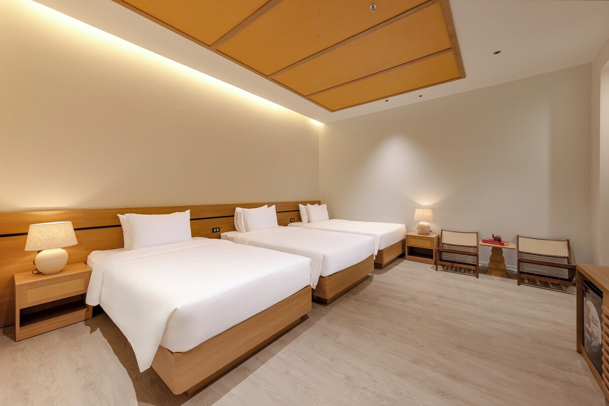 Habitación de hotel con dos camas, cabeceros de madera, mesitas de noche con lámparas, una pequeña mesa con sillas y un escritorio de madera.