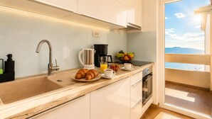 A modern kitchen with a sink, coffee maker, croissants, fruit bowl, and a view of the sea.