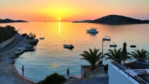 A coastal sunset view with boats, a promenade, and a hill in the background.