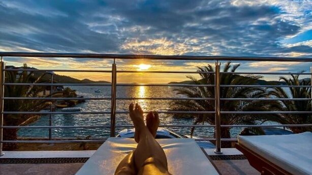 A balcony with a view of the sunset over the sea, a lounge chair, and palm trees.