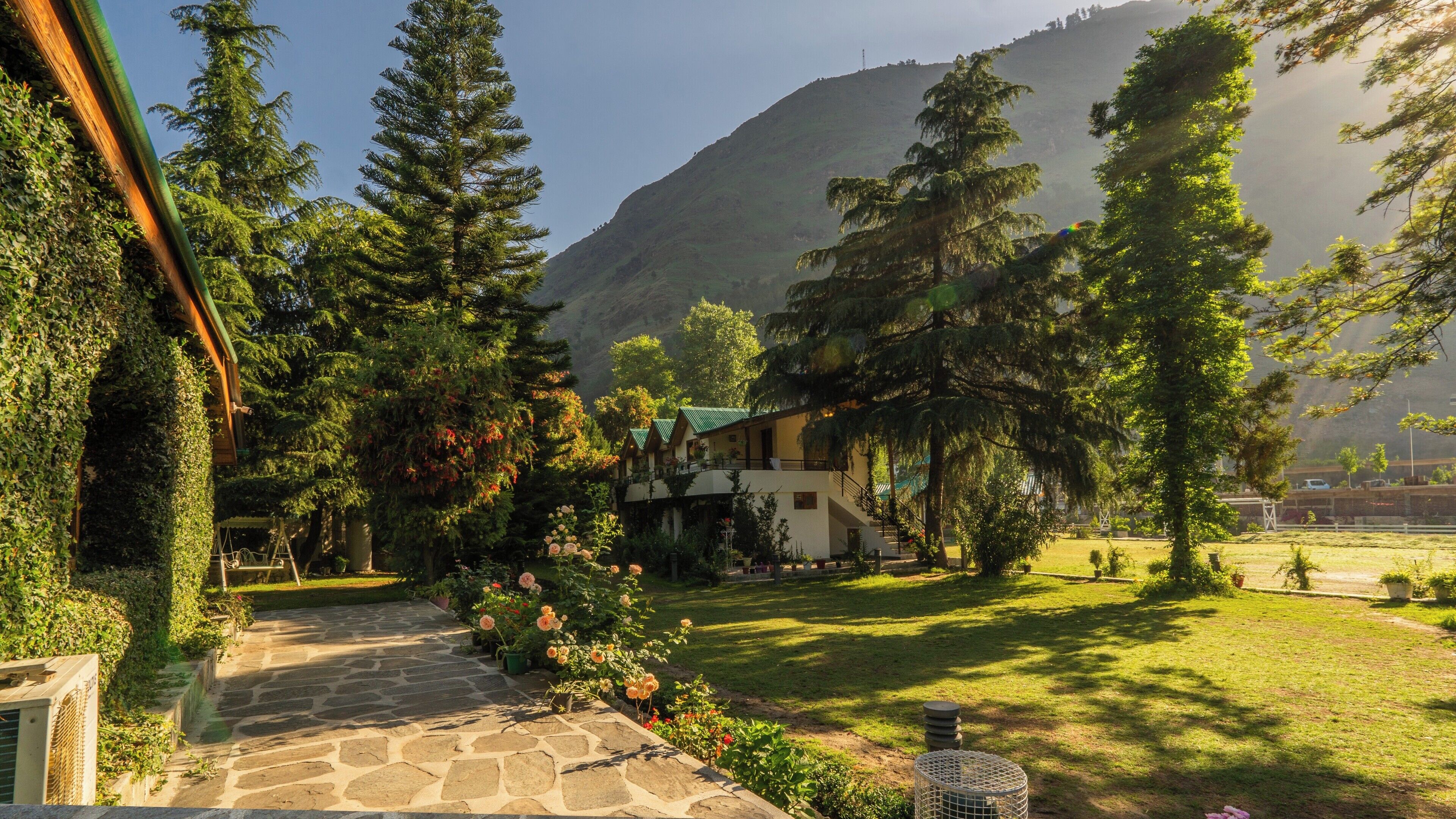 A mountain lodge with stone pathways, lush greenery, and a view of the surrounding hills.