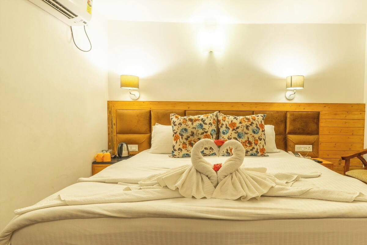 A hotel room with a bed, floral pillows, a heart-shaped towel decoration, a wooden headboard, bedside lamps, a wooden chair, and a window with curtains.