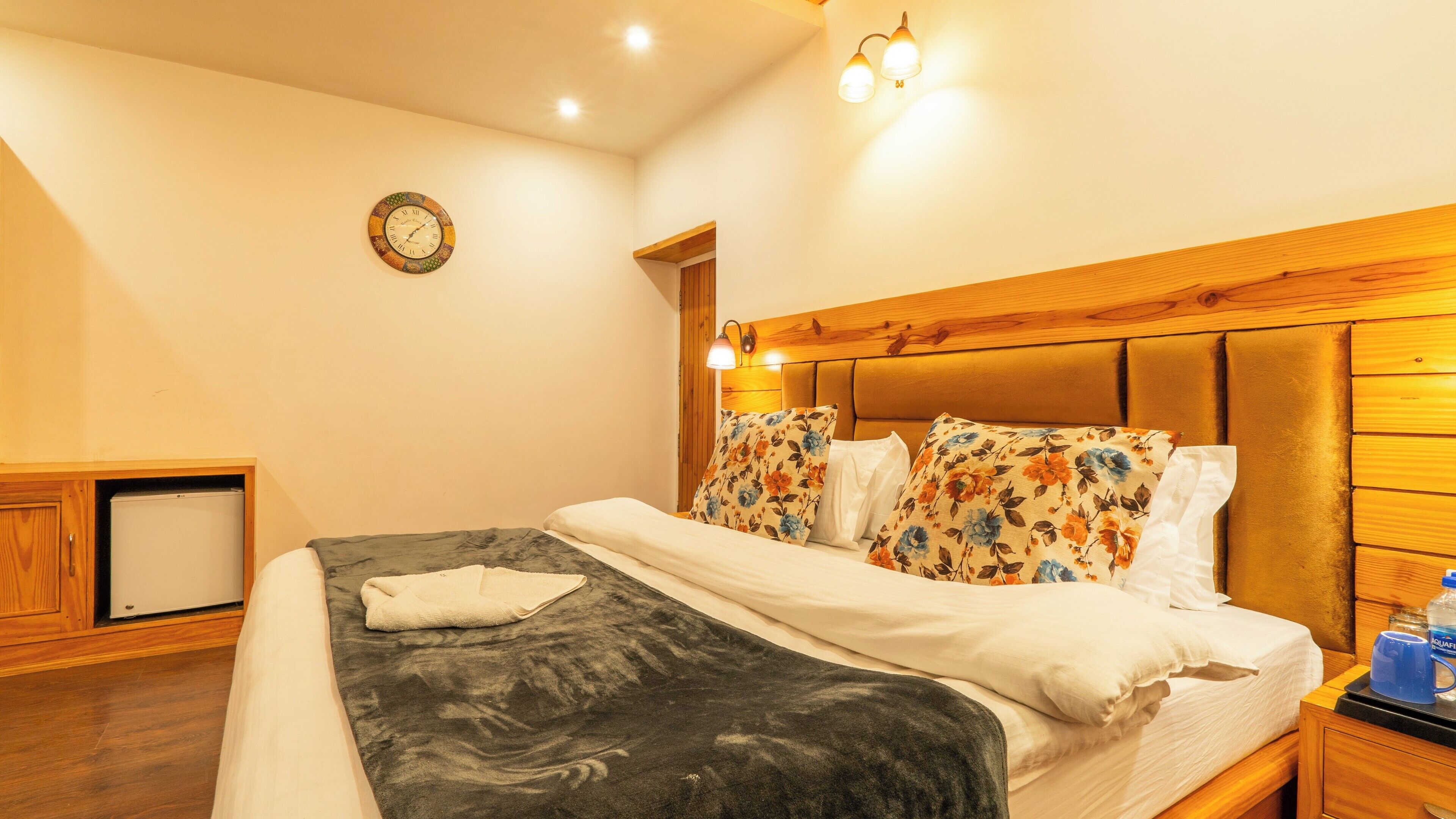 A hotel room with a bed, a nightstand, a wooden headboard, a wall clock, and a small desk with a lamp.