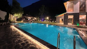 A swimming pool with a stone border and metal handrails, surrounded by a well-lit patio area with tables and chairs. - Smile Resort (Bhuntar)