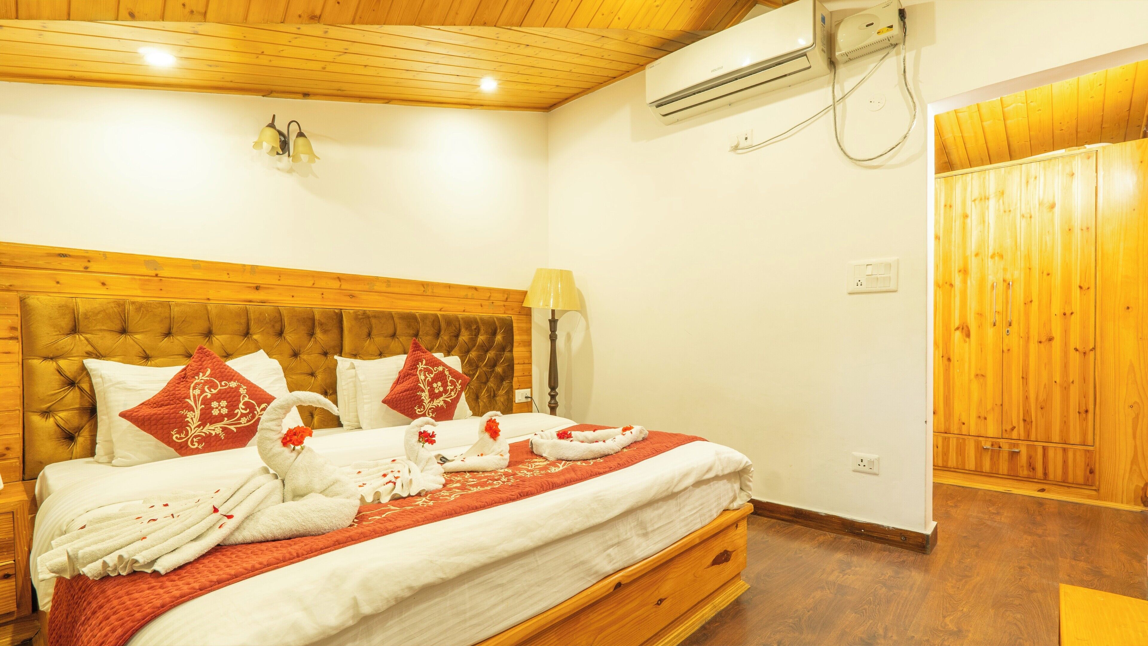Une chambre d’hôtel avec une tête de lit en bois, un lit avec une literie blanche et des oreillers rouges, une lampe de chevet et un climatiseur fixé au mur.