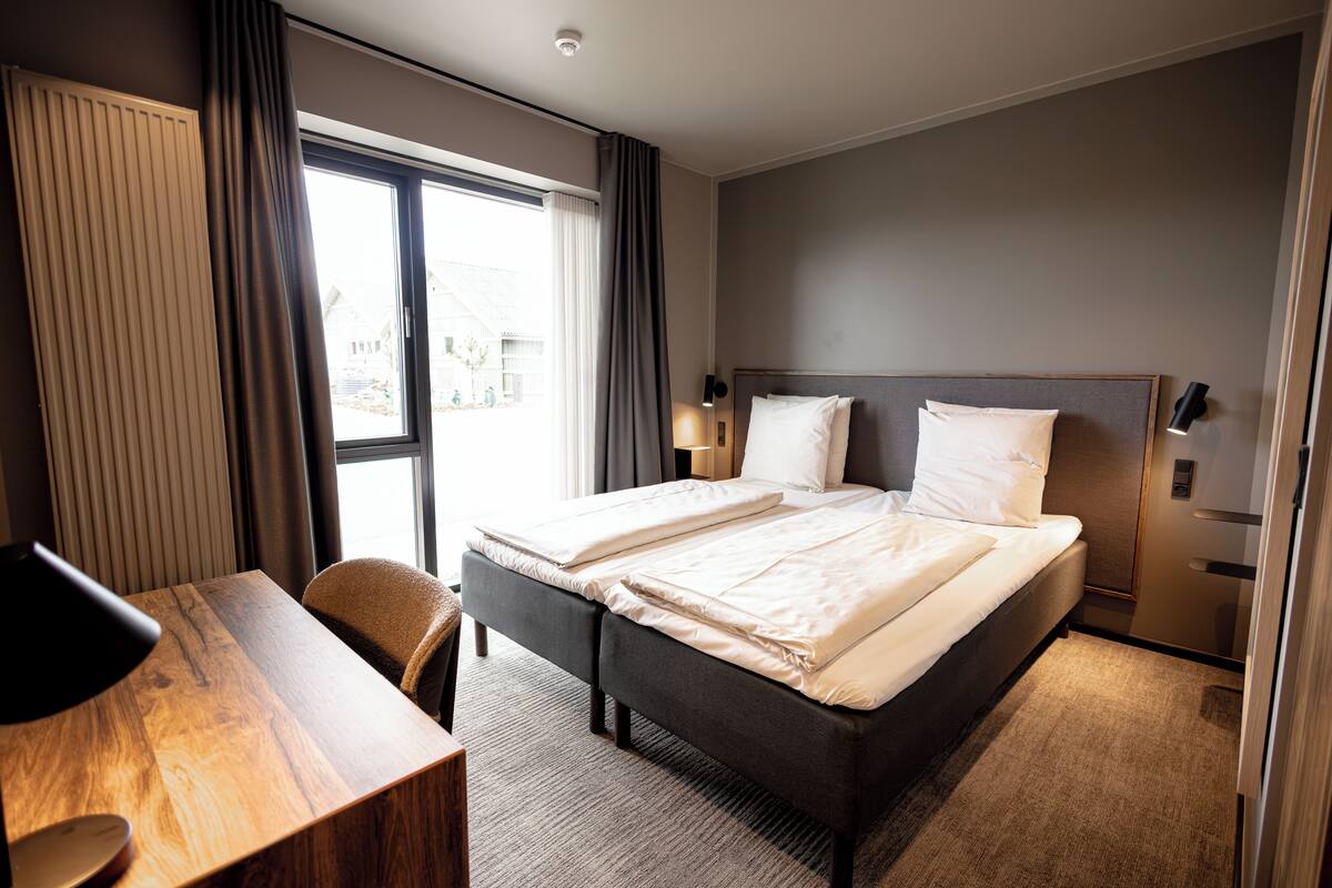 A hotel room with a bed, a desk, a chair, a lamp, and a window with curtains.