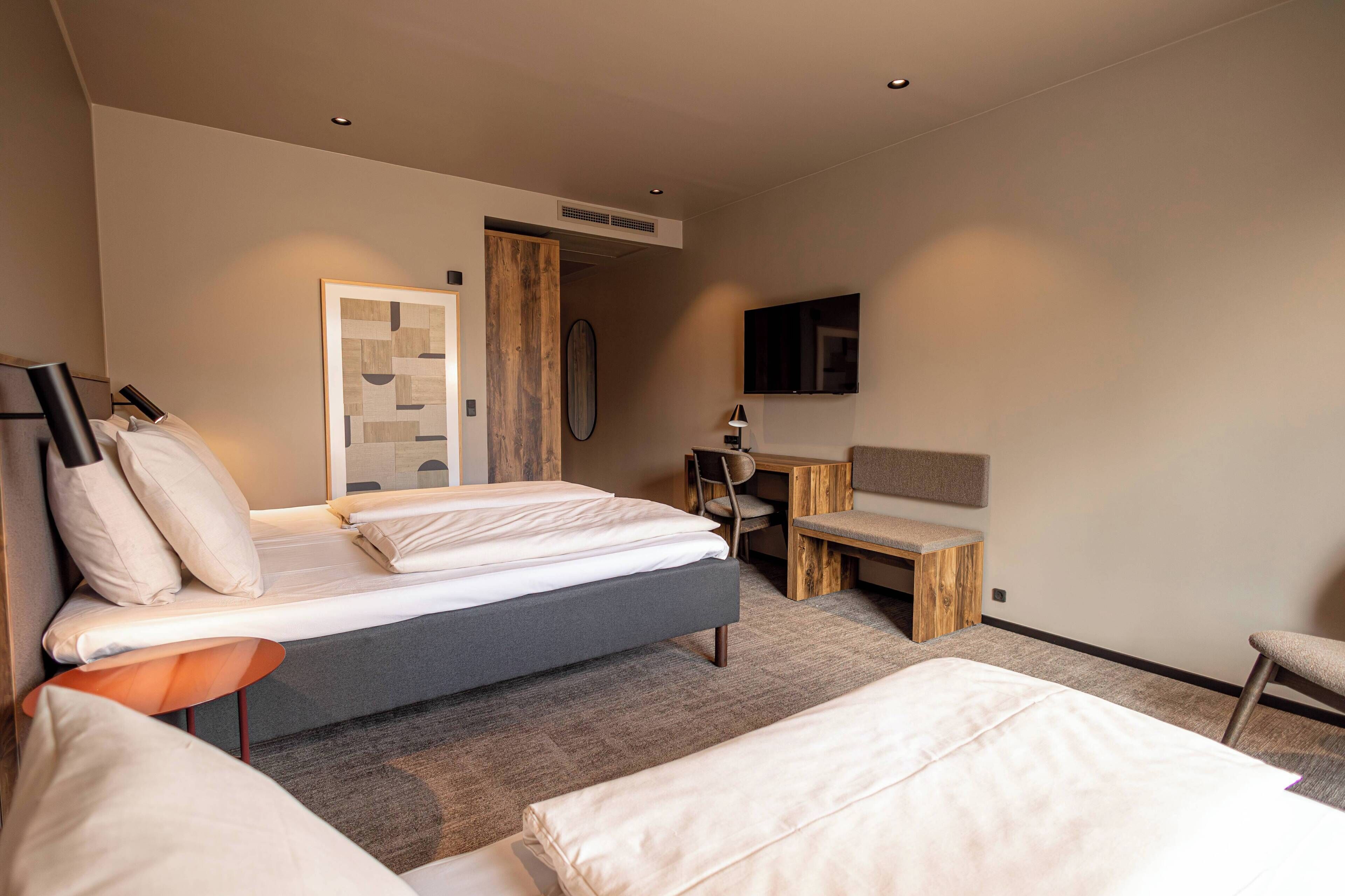 A modern hotel room with a bed, a desk, a chair, and a television on the wall.