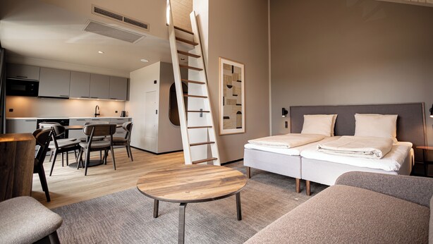 Une chambre d’hôtel moderne avec un lit en mezzanine, un coin repas avec une table et des chaises en bois, une cuisine en arrière-plan et un escalier menant à la mezzanine.