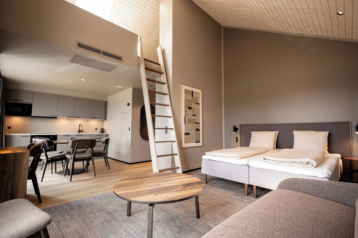 A modern hotel room with a loft bed, a dining area with a wooden table and chairs, a kitchen in the background, and a staircase leading to the loft.