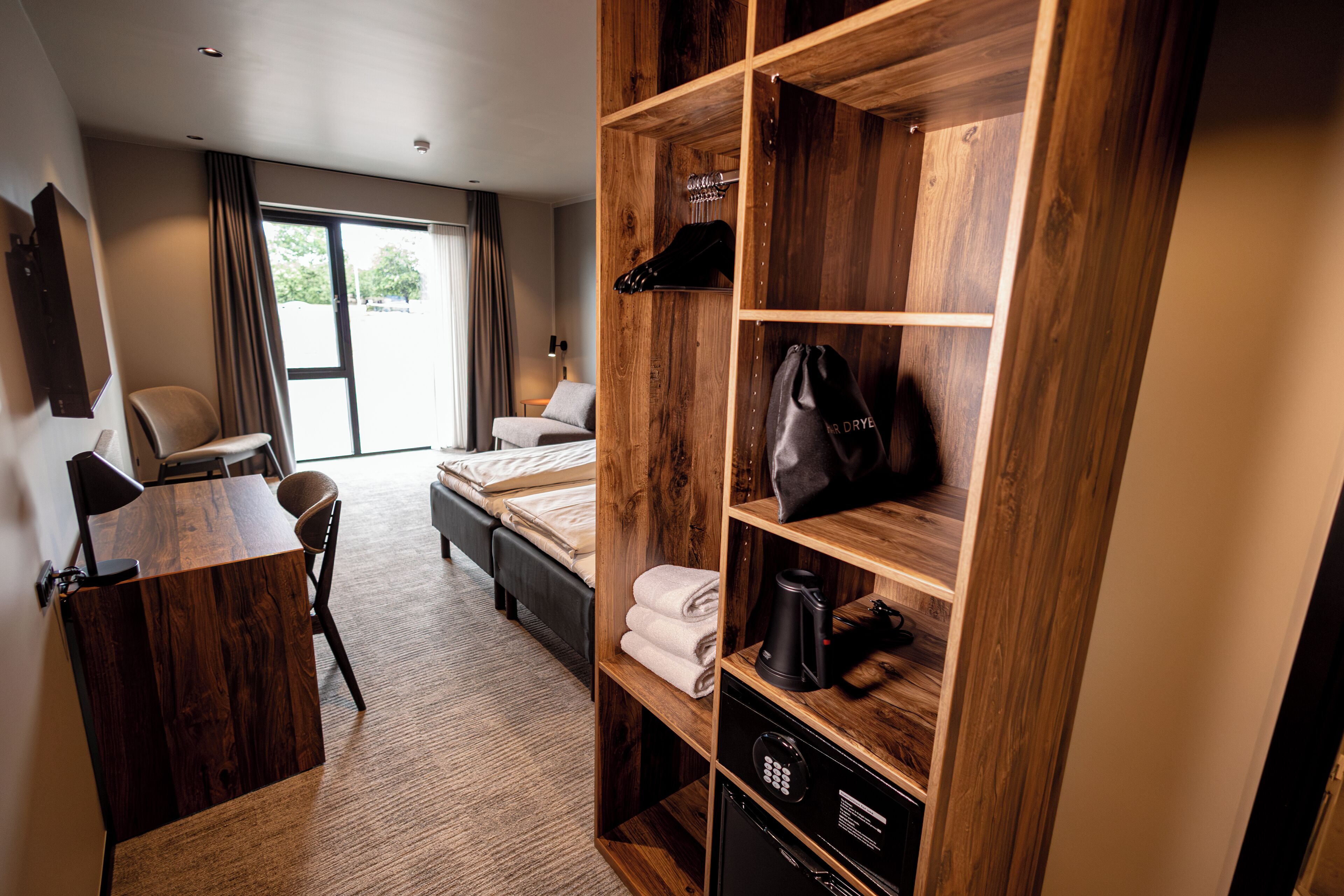 A hotel room with a wooden wardrobe, a desk, a bed, and a view of the outdoors.