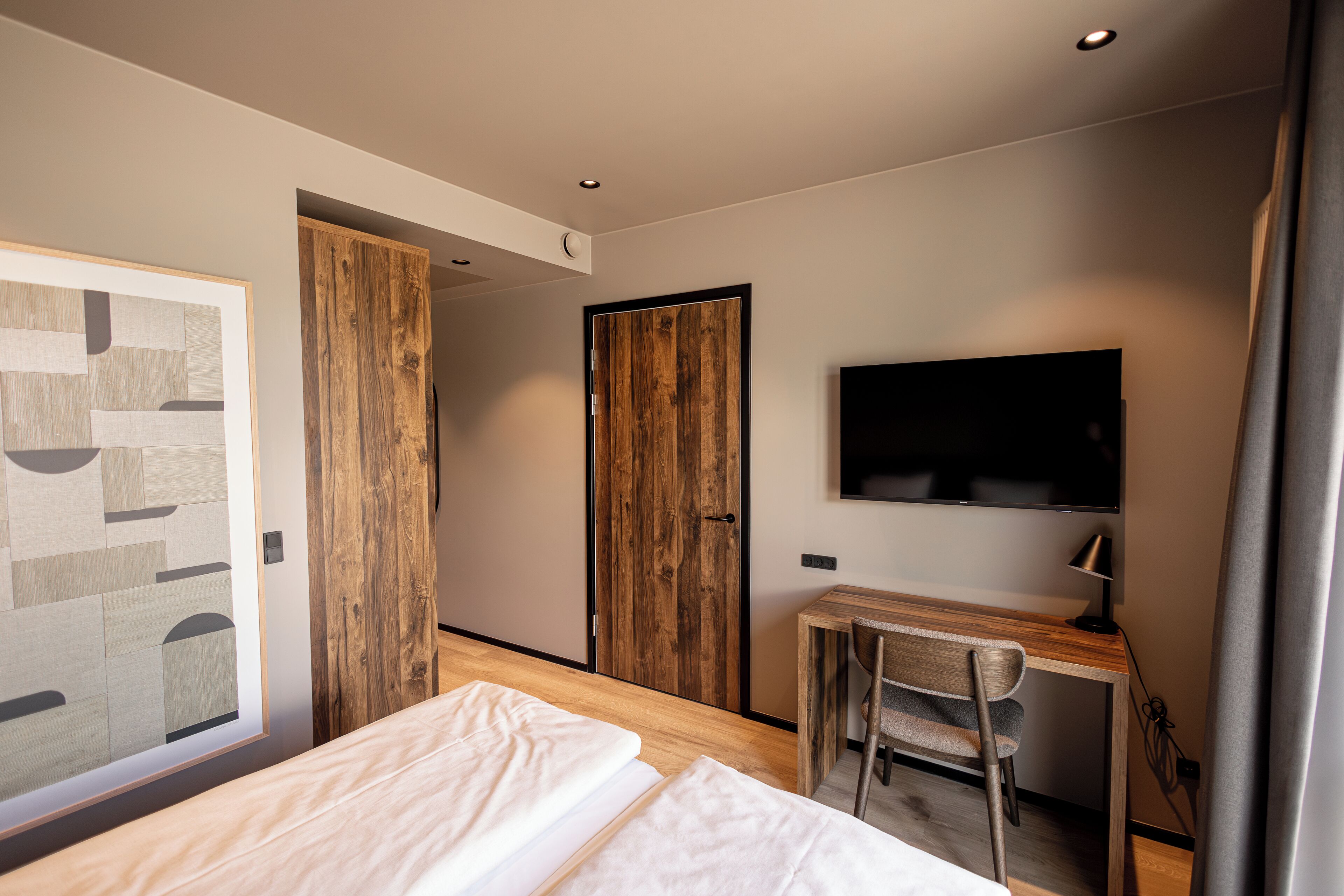 A modern hotel room with a wooden desk, a flat-screen TV, a bed with white linens, and a large window.