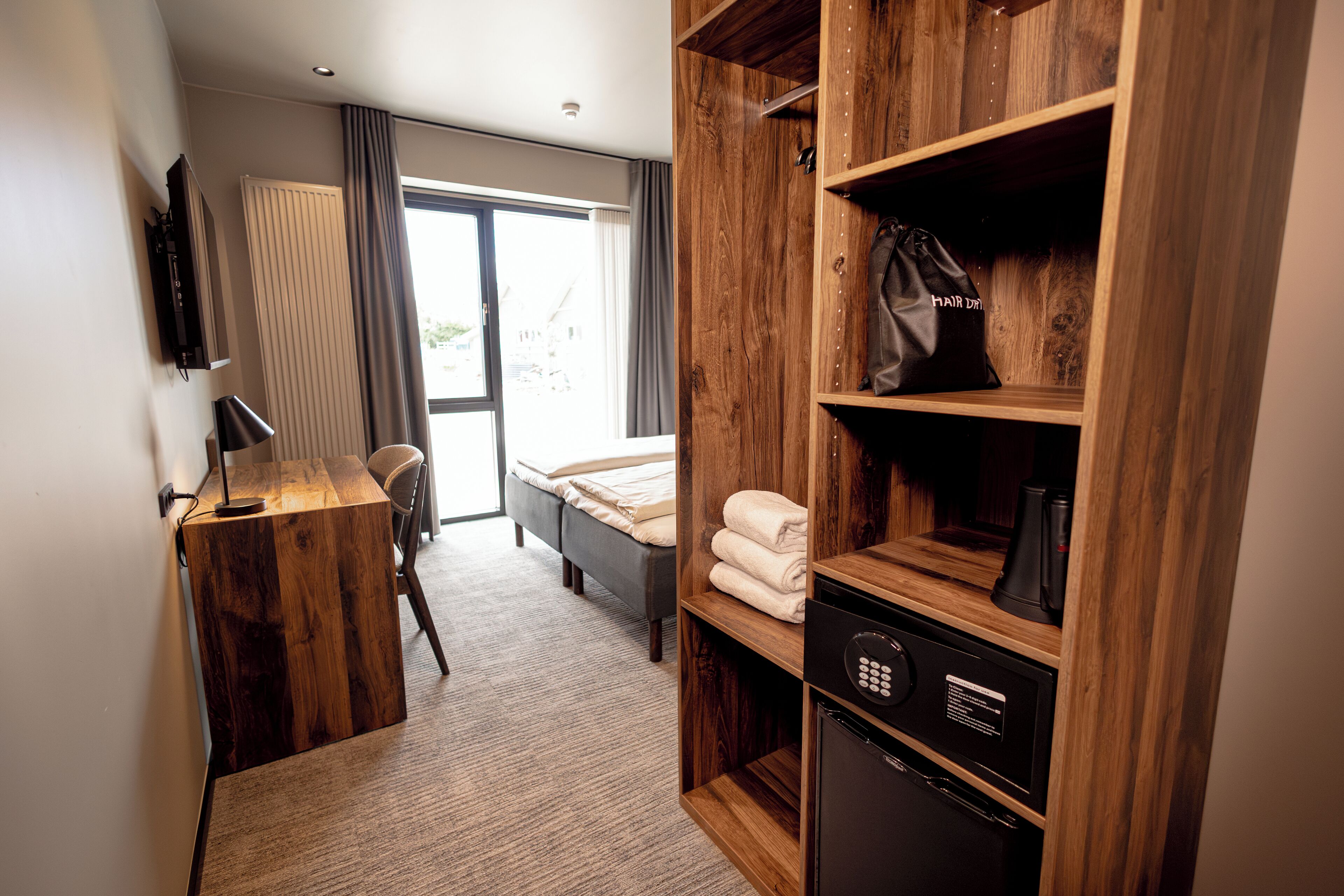 Habitación de hotel con un armario de madera, una cama, un escritorio con lámpara, televisión y vistas por una ventana.