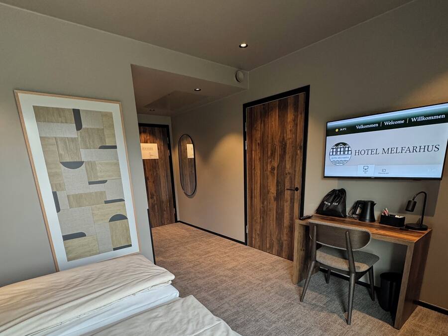 Habitación de hotel con cama, escritorio y televisor mostrando 'HOTEL MELFAHRUS'.