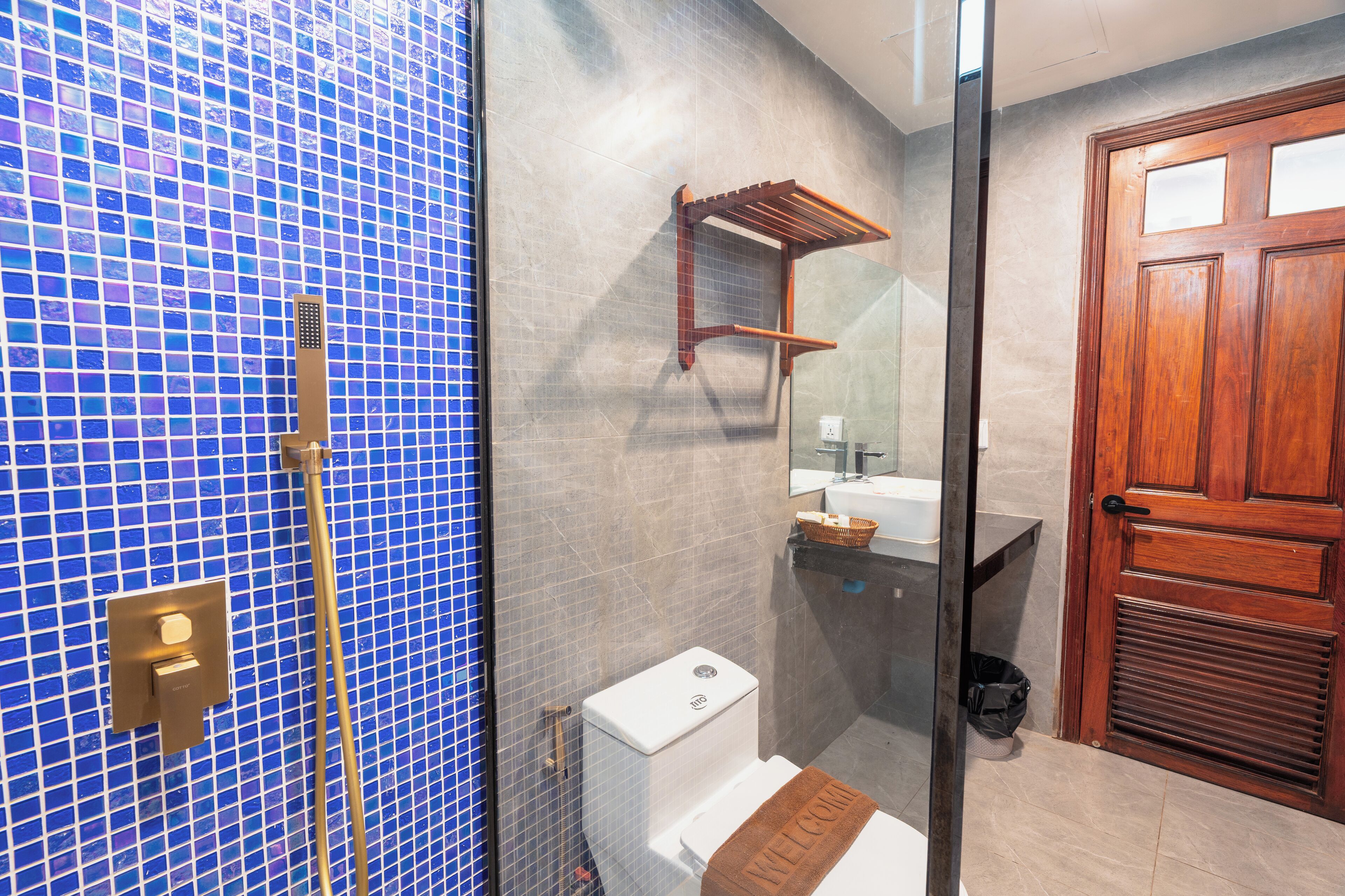 Un baño con pared de mosaicos azules, puerta de madera, inodoro, lavamanos con espejo y un perchero para toallas.