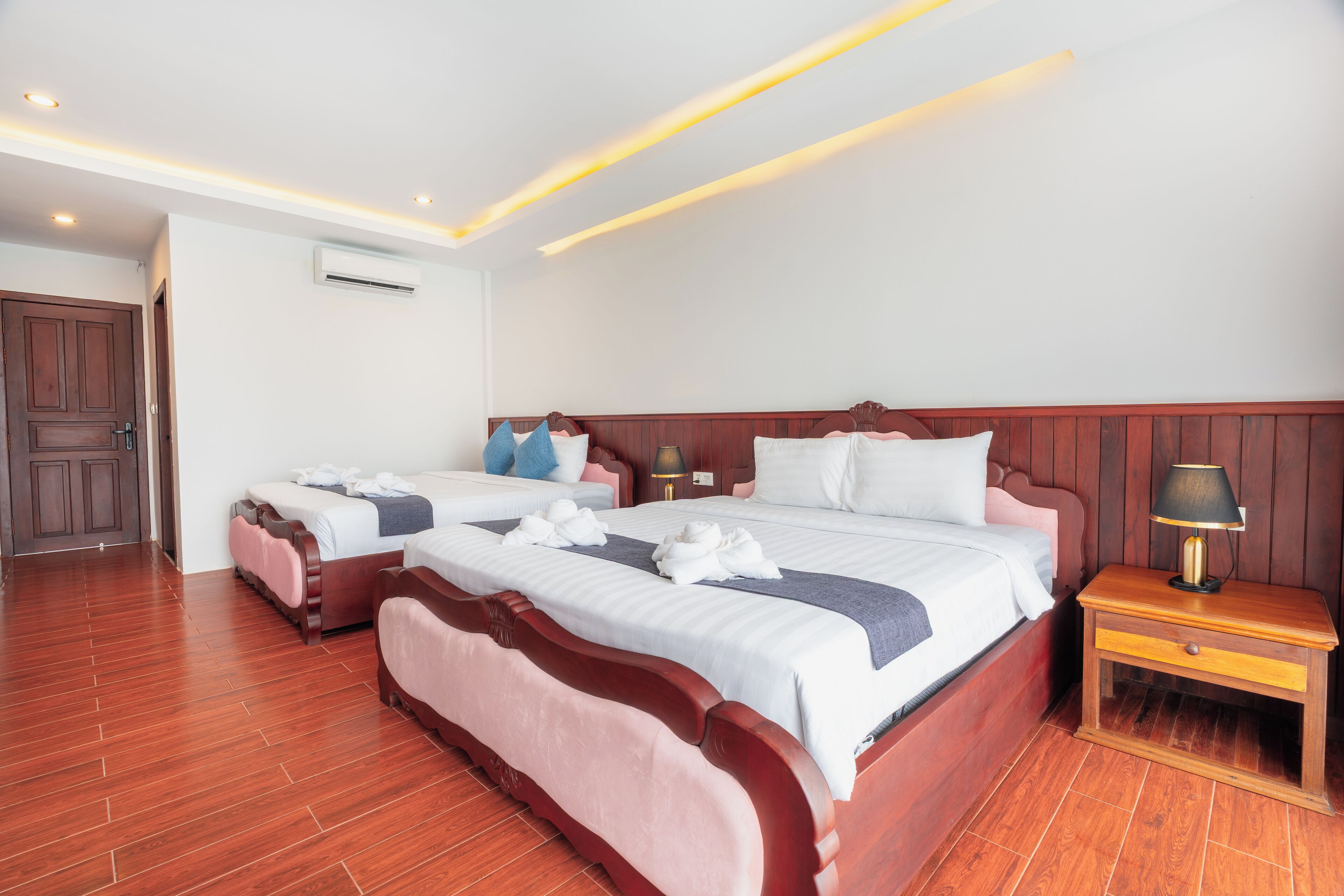 Habitación de hotel con dos camas, cabeceros de madera, una mesita de noche con lámpara y suelo de madera.