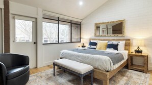 A bedroom with a large bed, a bench, a chair, a bedside table, and a mirror on the wall.