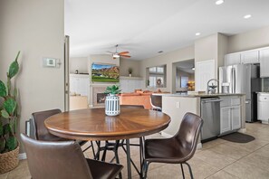 A modern kitchen with white cabinets, a stainless steel refrigerator, and a dining area with a wooden table and leather chairs. - La Quinta Desert Haven | Luxe Stay w/ Private Pool, Hot Tub, Pool Table (La Quinta)