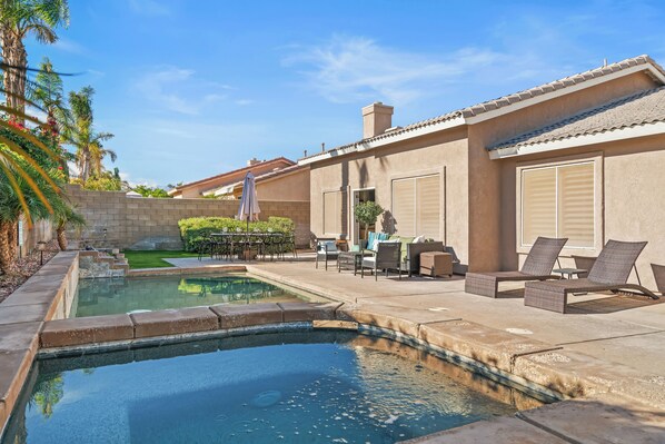 A swimming pool with a patio area, outdoor furniture, and a view of a building with large windows and a brick wall. - La Quinta Desert Haven | Luxe Stay w/ Private Pool, Hot Tub, Pool Table (La Quinta)