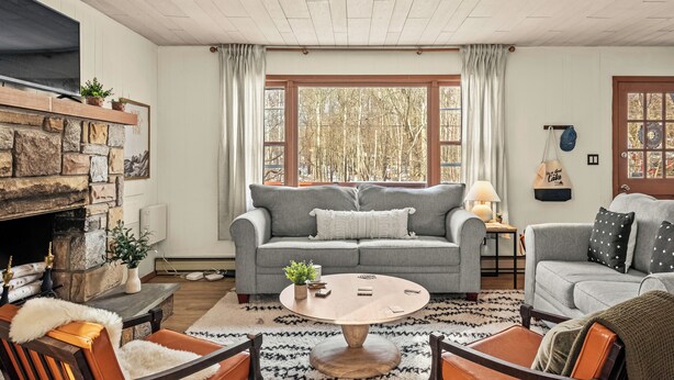 A cozy living room with a stone fireplace, grey sofas, a round coffee table, and a large window with curtains.