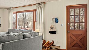A cozy living room with a gray sofa, a wooden door with glass panels, and a view of trees through the window.