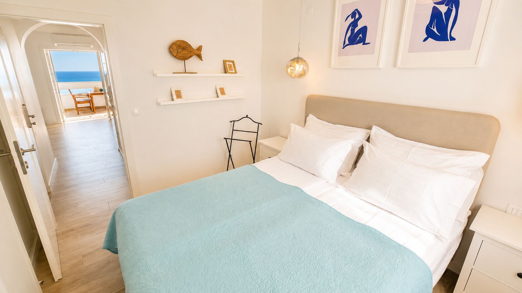 Ein Schlafzimmer mit einem Bett, einem Nachttisch, einer Ablagefläche, einem Regal mit Dekoration und Blick auf das Meer durch eine offene Tür.
