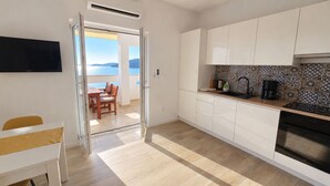 Una cocina moderna con armarios blancos, una mesa de comedor de madera y vista al mar a través de un ventanal abierto.