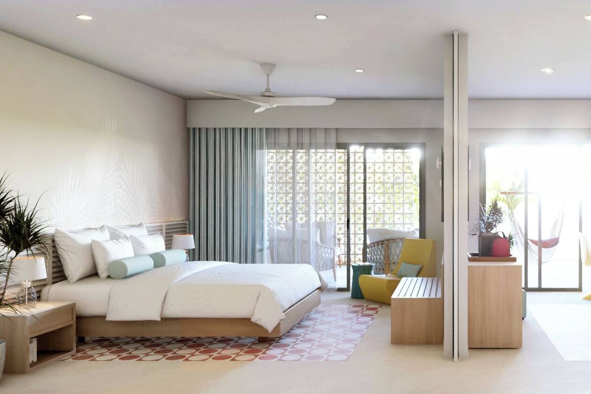 A modern hotel room with a bed, a sofa, a coffee table, and a chair. There is a large window with curtains and a view of the outdoors.