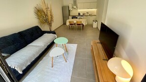 A compact living space with a sofa, two small tables, a TV, and a kitchen area in the background. - Eco-responsible haven of peace in Golfe-Juan: new serviced apartment (Vallauris)