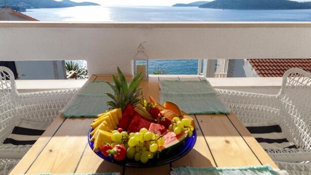 A table with a fruit platter and white wicker chairs with green cushions, overlooking a body of water and hills.