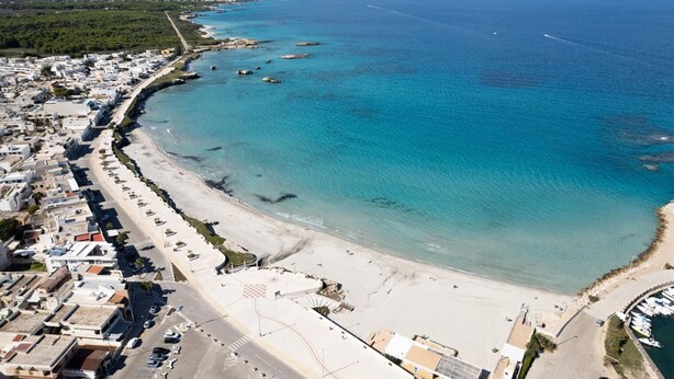 Vue aérienne d’une zone côtière comprenant une plage, des eaux d’un bleu limpide et une zone densément urbanisée avec des bâtiments et des voitures garées.
