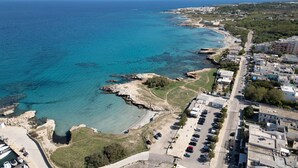 Vue aérienne d’une ville côtière, avec une mer bleue et limpide, une côte rocheuse et un parking rempli de voitures.