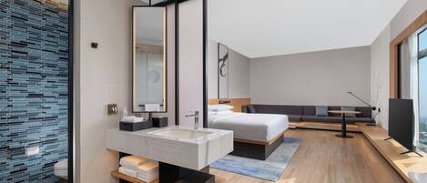 Un dormitorio moderno con una cama grande, una mesita de noche, un televisor y un baño con un espejo grande y lavamanos.