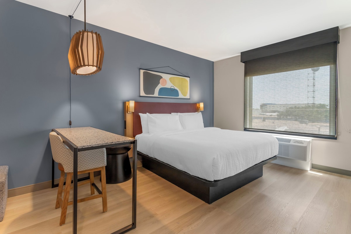 A modern hotel room with a bed, desk, chair, and a large window.