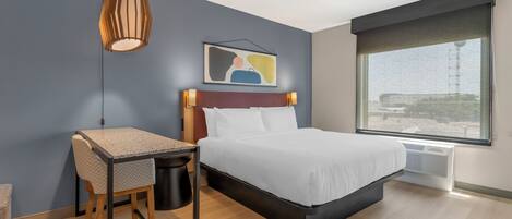 A modern hotel room with a bed, desk, chair, and a large window.