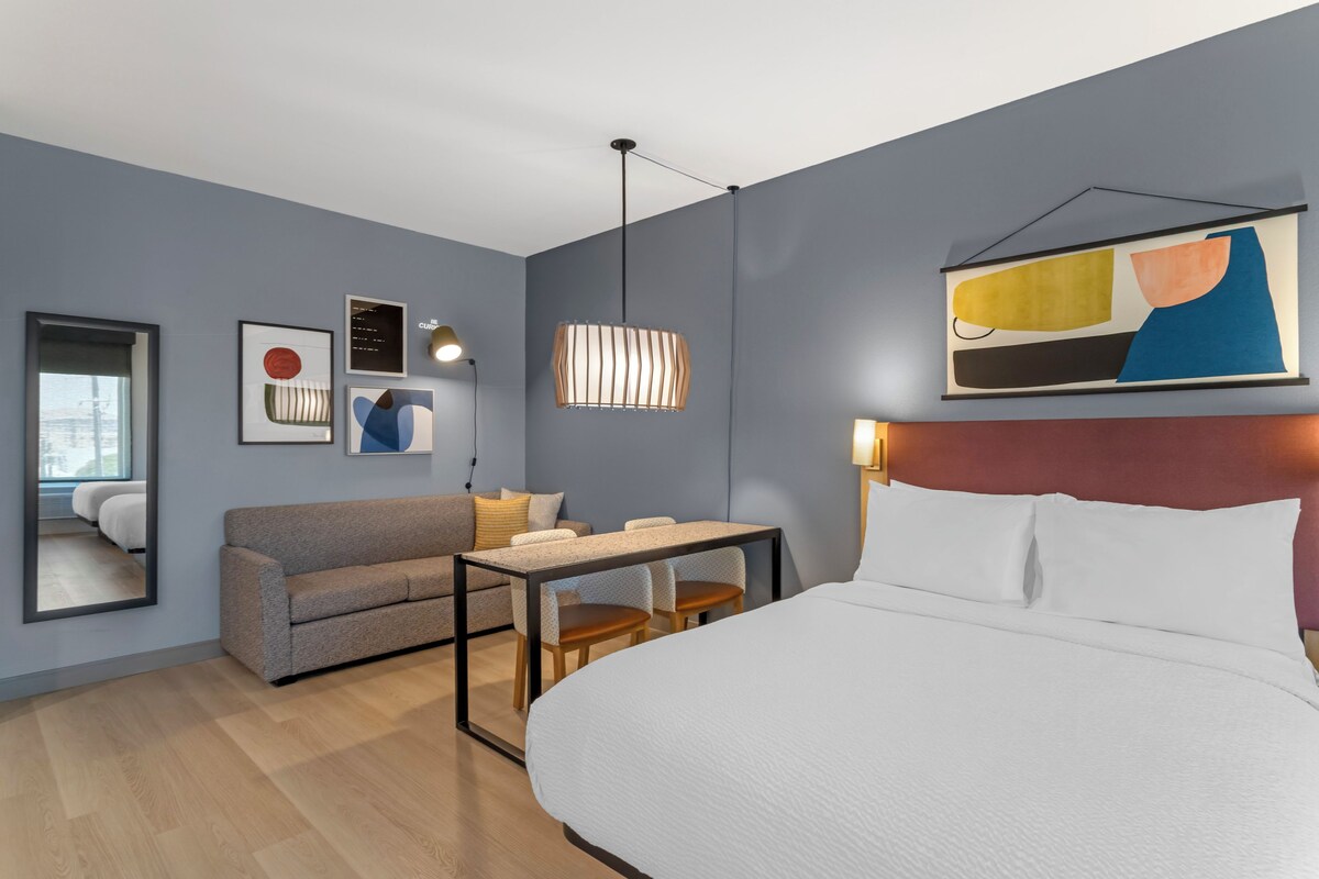 Et moderne hotelværelse med en stor seng, en sofa, et lille bord og et skrivebord. Væglamper og en loftslampe er monteret.