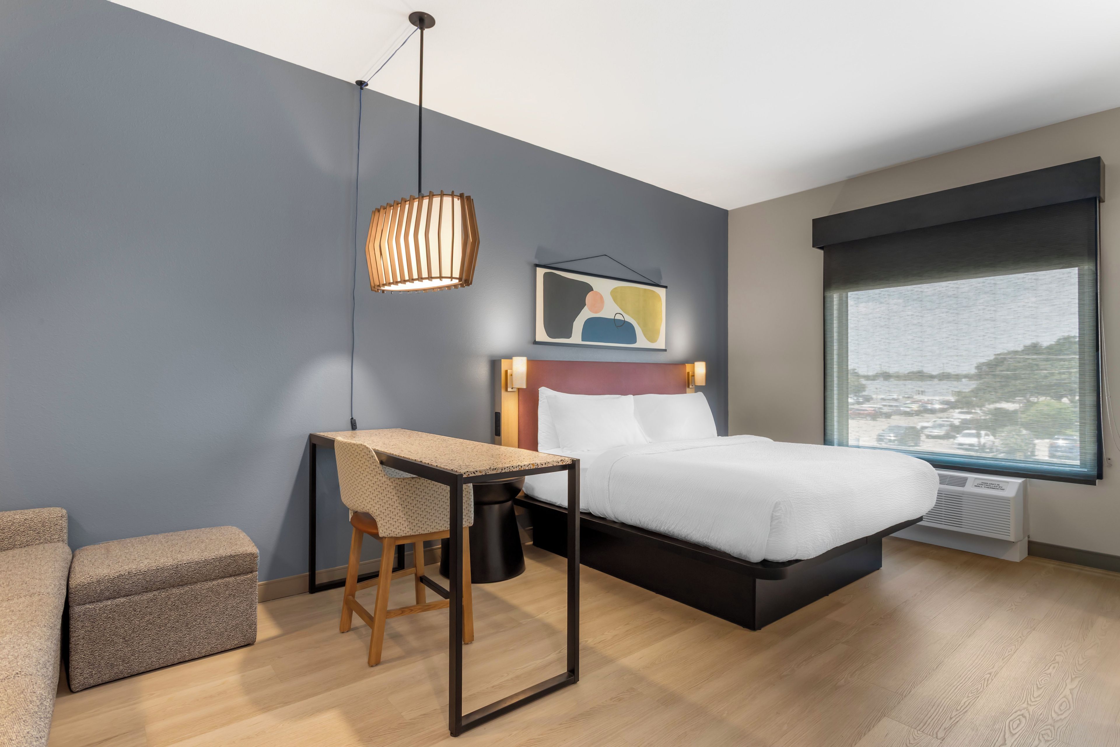 Una habitación de hotel moderna con cama, escritorio, silla, ventana grande y vista a la ciudad.