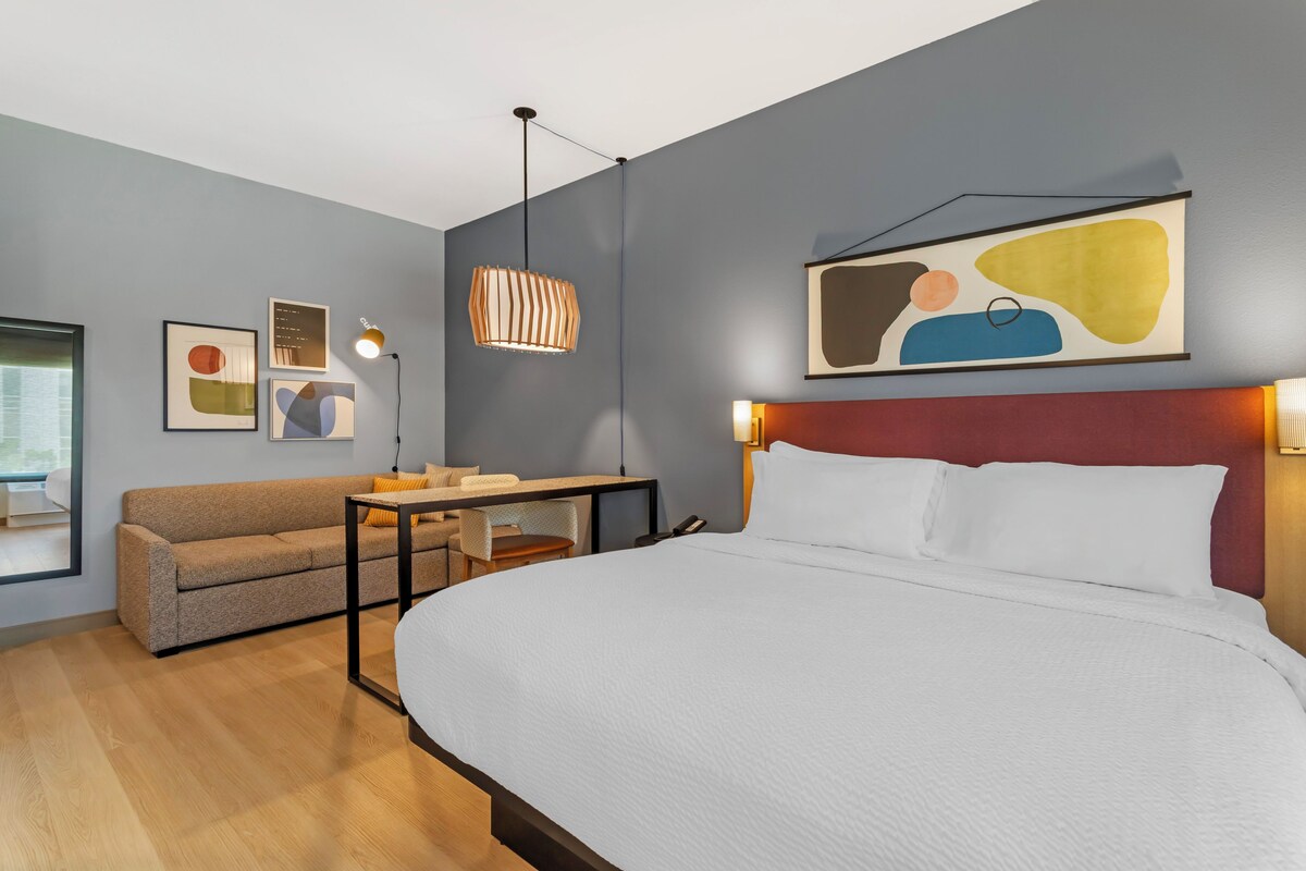 Une chambre d’hôtel moderne avec un grand lit, un canapé, une petite table et un bureau. Les murs sont ornés de tableaux abstraits et une fenêtre offre une vue agréable.