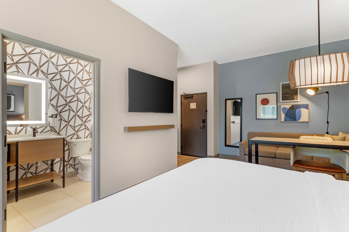 Et moderne hotelværelse med seng, skrivebord, spejl, fjernsyn og siddeområde med sofa og bord.