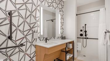 Un baño moderno con un revestimiento de pared con estampado geométrico, un lavamanos blanco con espejo, un mueble de tocador de madera y una ducha a ras de suelo con puerta de cristal.