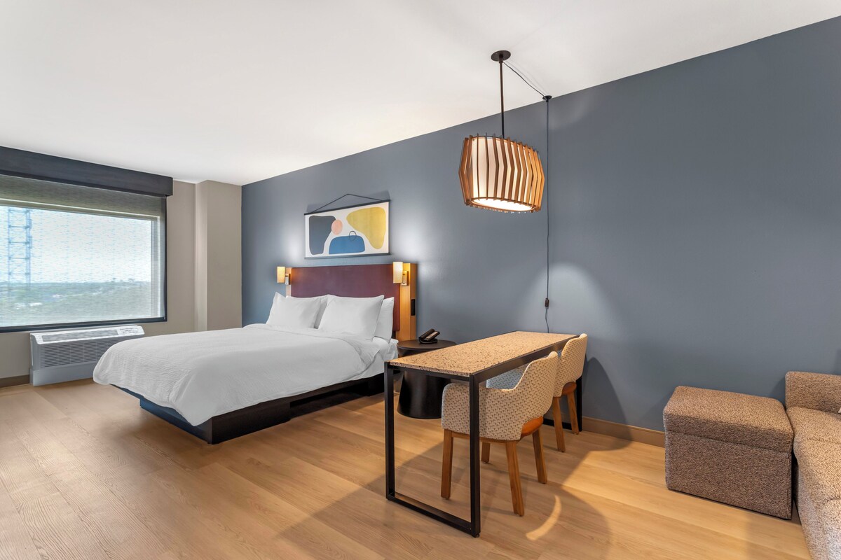 Et moderne hotelværelse med seng, skrivebord og stol, et stort vindue samt en markant vægmonteret lampe.