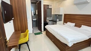 Habitación de hotel con cama, escritorio con silla, televisor, ventilador de techo y kitchenette.