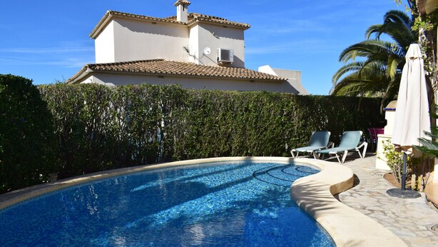 A swimming pool with a curved edge, surrounded by a stone patio. There are lounge chairs and an umbrella on the patio. A building with a tiled roof is visible in the background.
