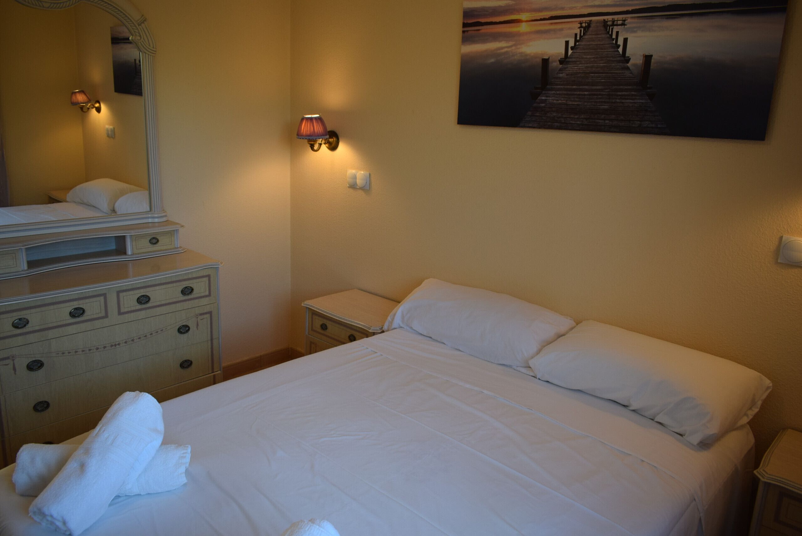 A hotel room with a bed, a dresser, a nightstand, a lamp, and a painting of a pier.
