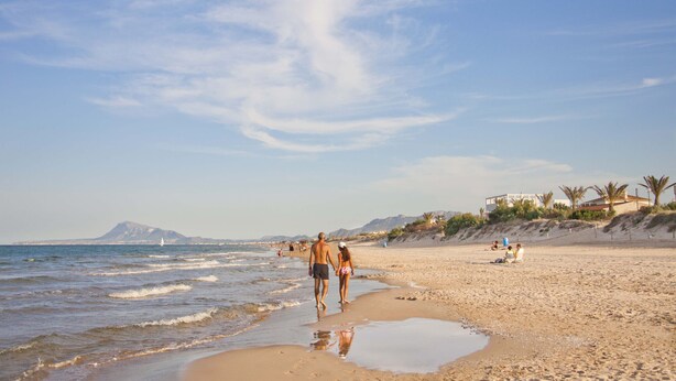Une plage où l’on voit des gens se promener le long du rivage, un voilier au loin et un bâtiment sur la droite.