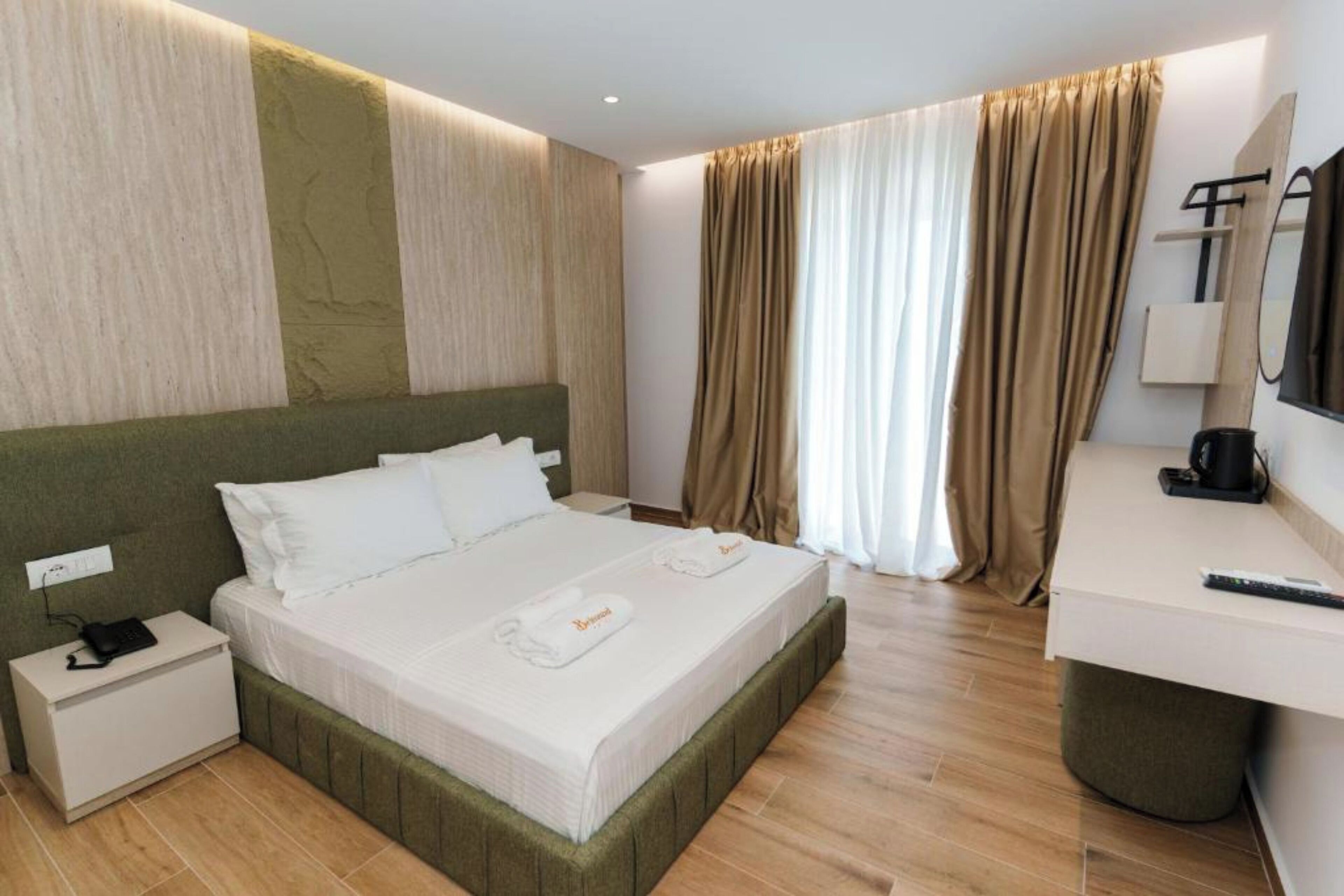 A hotel room with a large bed, a bedside table with a phone and charger, a desk with a coffee maker, and a mirror.