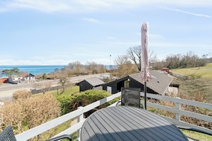 A deck with a view of the sea, a white fence, and a building with a dark roof.